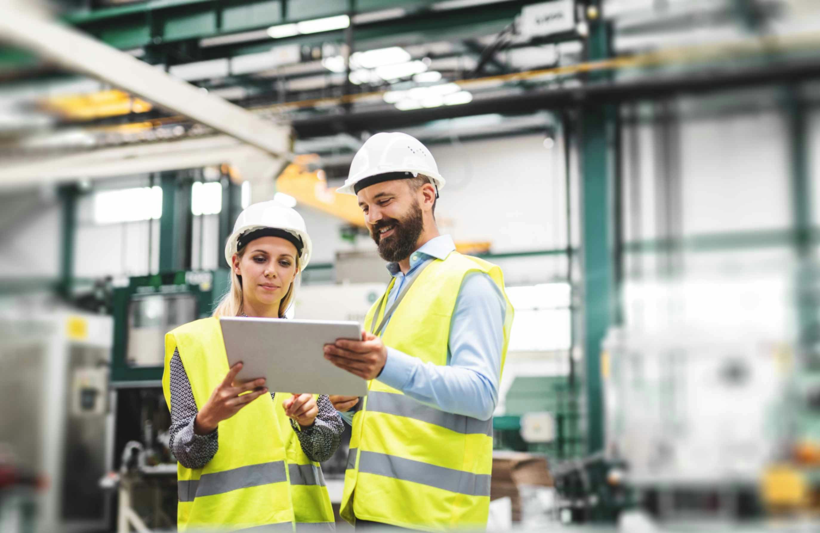 A man and a woman in hard hats and PPE looking at a tablet in a warehouse