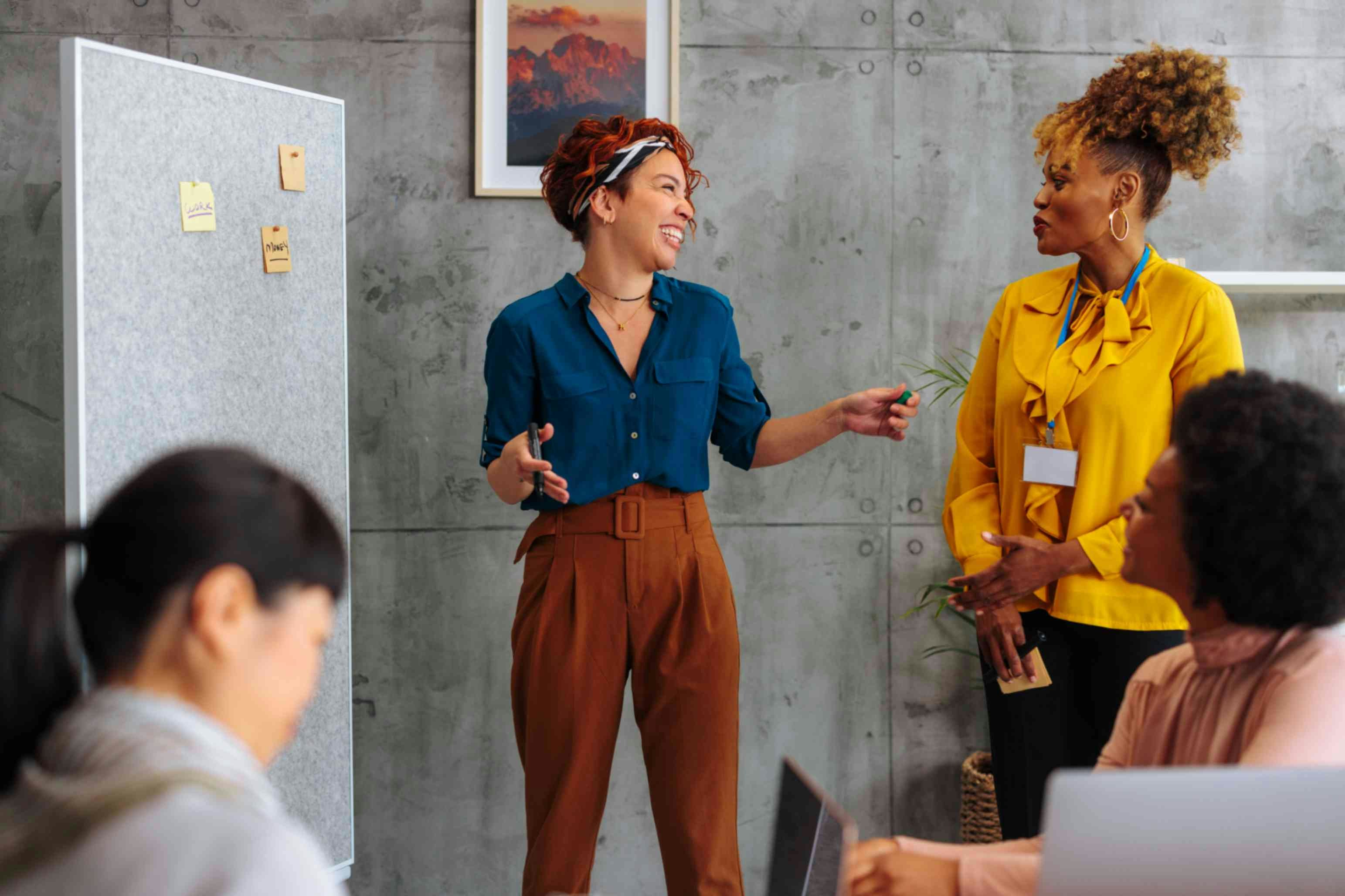 two women stood up giving a presentation, laughing in front of a note board and other women