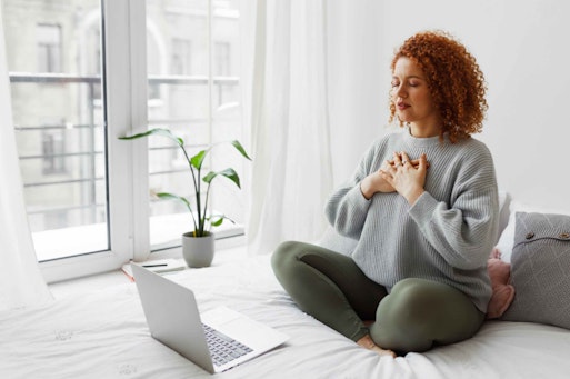 a woman sat on her bed in loungewear - with her hands to her heart - facing a computer.