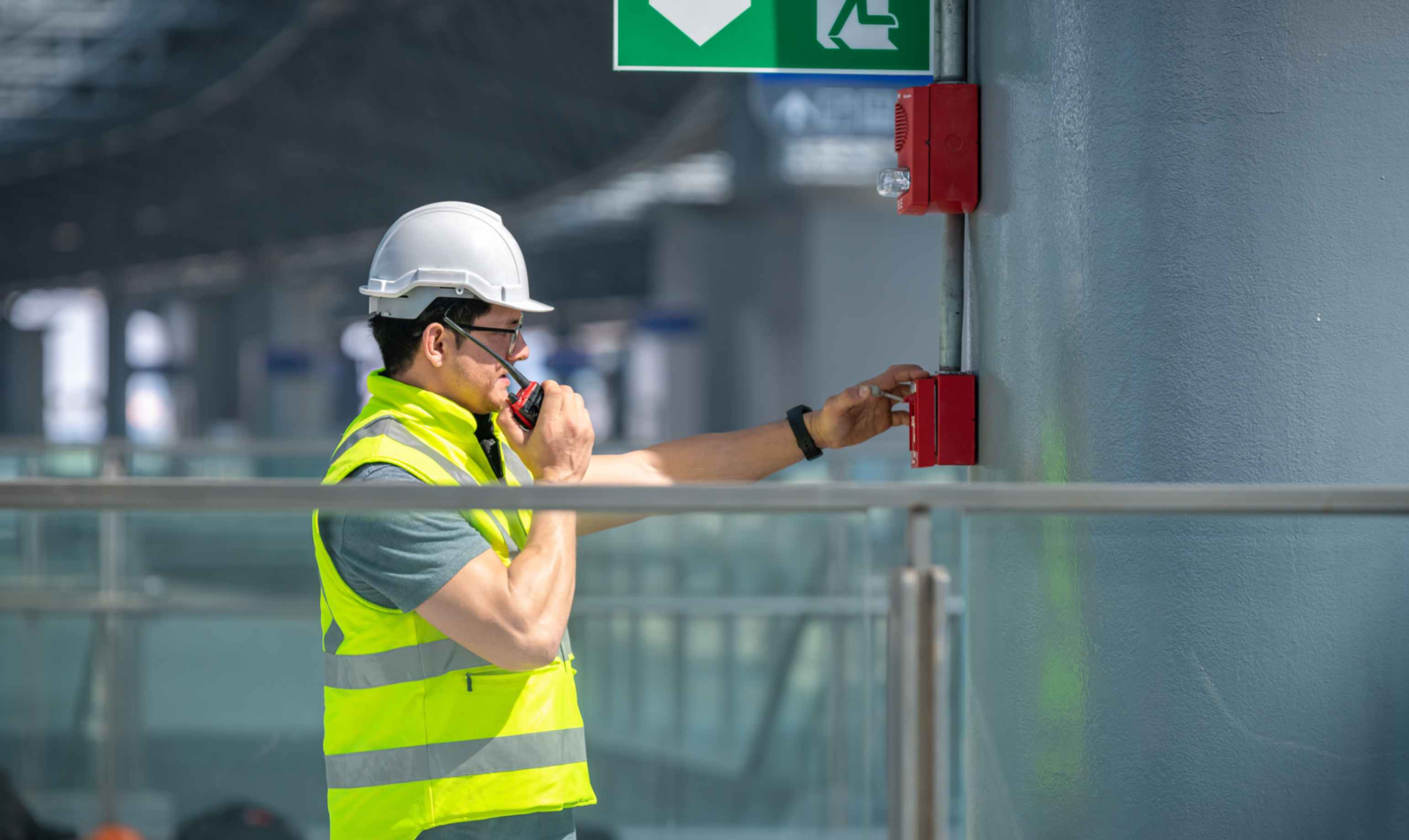 a man checking a fire alarm holding a walkie talkie and wearing PPE
