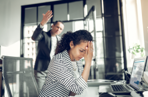 Employee with head in hands sat at desk. Other employer in background looks angry.