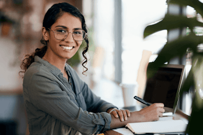 A smiling woman writing notes by her laptop.