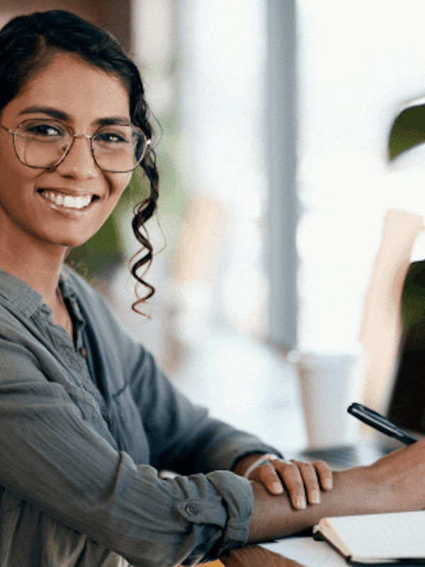 A smiling woman writing notes by her laptop.
