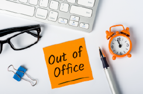 desk with out of office post it note, clock, pen and glasses around it