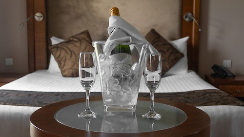 A bucket of champagne and two flutes in front of a bed, in a hotel room.