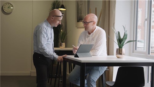Two man, one standing and one sat down, having a discussion next to a laptop.
