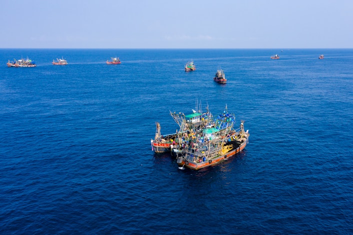 A photo of fishing vessels in the middle of the ocean, three of the vessels are tied to one another, there are eight others in the background.