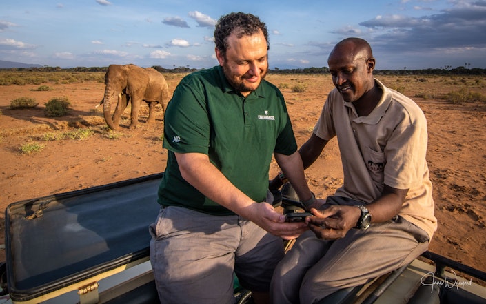 Jes Lefcourt and David Daballen sit in the back of an open top all terrain vehicle in Samburu, Kenya, as an elephant wanders in the background.