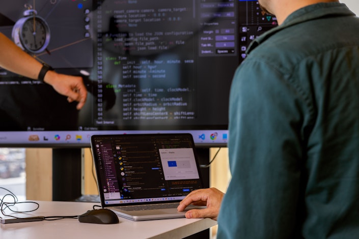 A closeup of someone using a laptop trackpad in front of a large monitor displaying code.
