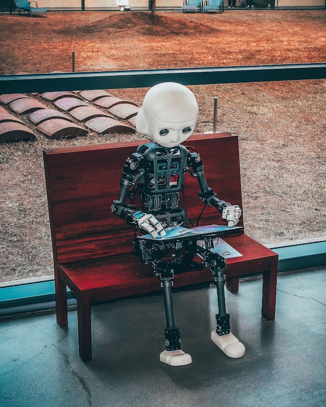 A robot sits on a wooden bench and reads a book.