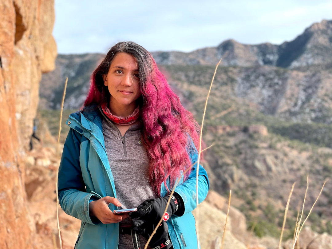 A photograph of Kiana, a woman with long hair dyed bright red, wearing a blue jacket and standing outside in front of mountains.