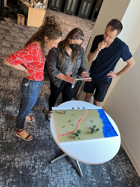 Three members of the hackathon team stand at a small table in an office. One member holds an iPad while the other two look on. On the table there is a small diorama featuring landscape.