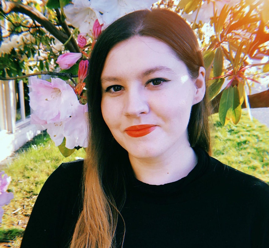 A photograph of Jenna James, a woman standing in front of a cherry blossom tree, wearing red lipstick and a black sweater.