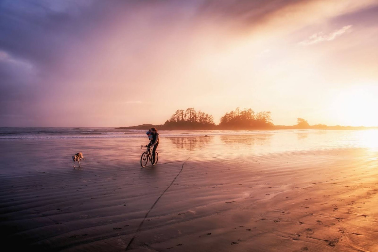 A man rides a bike on the beach while holding his son, with his golden retriever running alongside him.