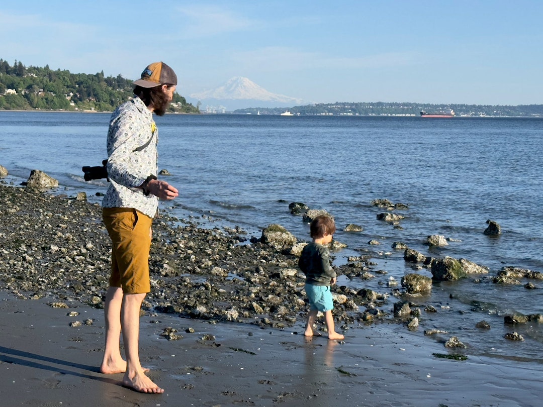 A man stands on a rocky beach, with his son standing in front of him.
