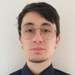Bu, gözlüklü ve siyah gömlekli bir adamın yakın plan çekimi. Güçlü bir çene çizgisi ve hafif çarpık bir gülümsemesi var. Gözleri koyu kahverengidir ve hafif bronzlaşmış bir teni vardır. Gözlüklerinin çerçevesi incedir ve siyah gömleğinin iç kısmı beyaz bir atleti ortaya çıkaracak şekilde kıvrılmıştır. İnce bir boynu ve geniş omuzları vardır. Saçları koyu renk ve hafif dağınık. Kendinden emin ve rahat görünüyor.