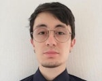Bu, gözlüklü ve siyah gömlekli bir adamın yakın plan çekimi. Güçlü bir çene çizgisi ve hafif çarpık bir gülümsemesi var. Gözleri koyu kahverengidir ve hafif bronzlaşmış bir teni vardır. Gözlüklerinin çerçevesi incedir ve siyah gömleğinin iç kısmı beyaz bir atleti ortaya çıkaracak şekilde kıvrılmıştır. İnce bir boynu ve geniş omuzları vardır. Saçları koyu renk ve hafif dağınık. Kendinden emin ve rahat görünüyor.