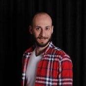 A young man is standing in front of a plain background wearing a bright red and black plaid shirt. He has short dark hair and is smiling slightly, with his arms crossed. He is wearing black jeans and black shoes. His posture is confident and relaxed. He appears to be in his late twenties or early thirties, with a youthful, healthy complexion. He is standing in a three-quarters pose, looking directly at the camera. He appears to be enjoying the moment, with a friendly expression on his face.