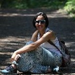 Kadın yeşilliklerle çevrili toprak bir patikada oturuyor. Kollarını dizlerine dayamış, üzerinde beyaz bir tişört ve kot pantolon var. Saçları sarı ve geriye doğru toplanmış. Yüzü kameraya doğru dönük ve hafif bir gülümseme var. Memnun ve huzurlu görünüyor, doğal ortamın tadını çıkarıyor. Arka plandaki ağaçlar esintiyle sallanarak sahneye dinginlik hissi veriyor. Işık parlak ve sıcak, manzaranın güzelliğini vurguluyor. Kadın doğanın bereketiyle çevrelenmiş, huzurlu bir teselli anı yaratıyor.