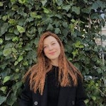 Genç bir kadın, yemyeşil yaprakları olan yemyeşil bir bitkinin önünde duruyor. Beyaz bir elbise ve siyah sandaletler giymiş, uzun siyah saçlarını at kuyruğu yapmış. Işıl ışıl gülümsüyor, ellerini kalçalarına dayamış bitkiye bakıyor. Güneş yüzünde ışıl ışıl parlıyor ve bitkinin parlak renkleri sahneye canlı ve neşeli bir hava katıyor. Bitki uzun ve canlı duruyor, yaprakları arka plandaki mavi gökyüzüyle canlı bir kontrast oluşturuyor.