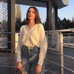 Bu görüntüde gündelik kıyafetiyle dışarıda duran bir kadın görülüyor. Elinde bir el çantası var ve koyu renk pantolonla beyaz bir üst giymiş. Saçları kısa bir bob şeklinde şekillendirilmiş ve yüzü hafifçe kameraya dönük. Kameradan uzağa bakıyor gibi görünüyor, ifadesi nötr ve dudakları hafifçe ayrılmış. Açık alanda tek başına dururken düşüncelere dalmış gibi görünüyor.