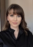 Bu, uzun, dalgalı kahverengi saçlı bir kadının görüntüsüdür. Uzun kollu ve yuvarlak yakalı siyah bir gömlek giyiyor. Yüz ifadesi sakin ve rahattır ve gözleri uzaklara odaklanmıştır. Küçük bir burnu var ve dudakları sanki bir şey söylemek üzereymiş gibi hafifçe ayrılmış. Saçları ortadan ayrılmış ve sırtından aşağı dökülüyor. Kendinden emin ve sakin görünüyor. Arka plan nötr bir renkte ve ışık yumuşak ve sıcak. Bu görüntü huzur ve dinginlik hissi veriyor.