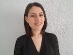 Sarı saçlı genç bir kadın kameraya mutlu bir şekilde gülümsüyor ve doğrudan objektife bakıyor. Açık ve dostça bir ifadesi var, gözleri geniş ve dudakları hafif bir sırıtış içinde. Yanakları hafifçe kızarmış ve teni ışıkta parlıyor. Beyaz bir tişört ve üzerinde küçük bir tılsım olan bir kolye takıyor. Saçları geriye doğru düşük bir at kuyruğu şeklinde toplanmış ve kolları göğsünün önünde kavuşturulmuş. Düz beyaz bir arka planın önünde duruyor ve yanında birkaç bitki var. Rahat ve memnun görünüyor, anı yaşıyor ve kamera önünde geçirdiği zamanın tadını çıkarıyor gibi görünüyor.