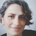 Uzun kahverengi saçlı, beyaz tişört ve mavi kot pantolon giyen neşeli bir kadın beyaz bir arka planın önünde duruyor ve kameraya gülümsüyor. Gözleri parlak, dudakları hafifçe yukarı doğru kıvrılmış, yüzünde sıcak ve davetkâr bir ifade var. Ellerini önünde tutmuş, sanki beklenti içindeymiş gibi birbirine kenetlemiş. Gülümsemesi samimi ve tüm tavırları davetkâr. Kendinden emin ve memnun görünüyor, neşe ve mutluluk hissi yayıyor. Gururlu ve güçlü bir duruşa sahip, başı dik ve bakışları kendinden emin.