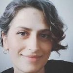 Uzun kahverengi saçlı, beyaz tişört ve mavi kot pantolon giyen neşeli bir kadın beyaz bir arka planın önünde duruyor ve kameraya gülümsüyor. Gözleri parlak, dudakları hafifçe yukarı doğru kıvrılmış, yüzünde sıcak ve davetkâr bir ifade var. Ellerini önünde tutmuş, sanki beklenti içindeymiş gibi birbirine kenetlemiş. Gülümsemesi samimi ve tüm tavırları davetkâr. Kendinden emin ve memnun görünüyor, neşe ve mutluluk hissi yayıyor. Gururlu ve güçlü bir duruşa sahip, başı dik ve bakışları kendinden emin.
