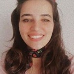 Uzun kumral saçlı genç bir kadın doğrudan kameraya gülümsüyor. Yumuşak yüz hatları ve sıcak bir ifadesi var. Gözleri parlak ve dudakları rahat bir gülümsemeyle hafifçe ayrılmış. Kısa kabarık kollu ve beyaz yakalı soluk pembe bir üst giyiyor. Elleri vücudunun önünde birbirine kenetlenmiş. Yanakları pembe ve yüzü uzun saçlarıyla çerçevelenmiş. Doğrudan kamera objektifine bakarken eğleniyor gibi görünüyor. Arka plan parlak ve havadar, biraz da mavi. Kadın mutlu, memnun ve kendinden emin görünüyor ve neşe hissi yayıyor.