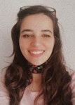 Uzun kumral saçlı genç bir kadın doğrudan kameraya gülümsüyor. Yumuşak yüz hatları ve sıcak bir ifadesi var. Gözleri parlak ve dudakları rahat bir gülümsemeyle hafifçe ayrılmış. Kısa kabarık kollu ve beyaz yakalı soluk pembe bir üst giyiyor. Elleri vücudunun önünde birbirine kenetlenmiş. Yanakları pembe ve yüzü uzun saçlarıyla çerçevelenmiş. Doğrudan kamera objektifine bakarken eğleniyor gibi görünüyor. Arka plan parlak ve havadar, biraz da mavi. Kadın mutlu, memnun ve kendinden emin görünüyor ve neşe hissi yayıyor.