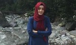 Boynuna parlak kırmızı bir başörtüsü bağlamış bir kadın kameraya bakıyor. Koyu renk gözleri uzun siyah kirpikleriyle çerçevelenmiş ve yüzünde ciddi bir ifade var. Hafif bir kumaştan yapılmış olan eşarp omuzlarından ve göğsünden aşağıya yumuşak bir şekilde düşüyor. Saçları yüzünden geriye doğru toplanmış ve kollarını önünde kavuşturmuş bir şekilde duruyor. Kendinden emin ve kararlı görünüyor, karşısına ne çıkarsa çıksın üstesinden gelmeye hazır.