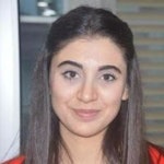 Bu, uzun, dalgalı kahverengi saçlı, parlak bir gülümseme takınmış genç bir kadının görüntüsüdür. Gözleri geniş ve pırıl pırıl, dudakları ise kıvrımlı bir neşe ifadesi. Açık tenli ve saçları ortadan ayrılmış. Üzerinde düz beyaz bir tişört var ve yanakları hafifçe kızarmış. Arka plan yumuşak, kirli beyaz bir renktedir. Kadın doğrudan kameraya bakıyor ve ifadesinde memnuniyet ve mutluluk var.