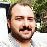Kalın, kara bıyıklı bir adam genişçe gülümsüyor, yüzü neşeyle aydınlanmış. Beyaz yakalı bir gömlek giymiş, üst düğmesi açık. Gözleri parlak ve köşeleri kırışık, dudakları neşeli bir sırıtışla kıvrılmış. Elleri göğsünün önünde birbirine kenetlenmiş ve sanki gülüyormuş gibi başı hafifçe arkaya eğilmiş. Arka plan bulanıklaştırılmış ve adam görüntünün odağı haline getirilmiş. Memnun ve kaygısız görünüyor.