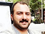 Kalın, kara bıyıklı bir adam genişçe gülümsüyor, yüzü neşeyle aydınlanmış. Beyaz yakalı bir gömlek giymiş, üst düğmesi açık. Gözleri parlak ve köşeleri kırışık, dudakları neşeli bir sırıtışla kıvrılmış. Elleri göğsünün önünde birbirine kenetlenmiş ve sanki gülüyormuş gibi başı hafifçe arkaya eğilmiş. Arka plan bulanıklaştırılmış ve adam görüntünün odağı haline getirilmiş. Memnun ve kaygısız görünüyor.