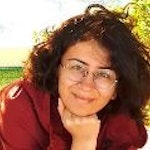 Bu, elini çenesine doğru kaldırmış, düşünceli bir ifadeyle uzaklara bakan bir kadının görüntüsüdür. Uzun, koyu renk saçları yüzünden geriye doğru toplanmış. Gözleri odaklanmış ve dudakları derin düşüncelere dalmış gibi hafifçe ayrılmış. Üzerinde beyaz bir bluz, siyah bir hırka ve ince altın bir kolye var. Beden dili rahat ve duruşu kendinden emin. Dikkatli ve düşünceli bir şekilde bir şeyler düşünüyor gibi görünüyor. Bakışları mesafeli ve yüzü sıcak, doğal bir ışıkla aydınlatılmış.