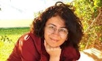 Bu, elini çenesine doğru kaldırmış, düşünceli bir ifadeyle uzaklara bakan bir kadının görüntüsüdür. Uzun, koyu renk saçları yüzünden geriye doğru toplanmış. Gözleri odaklanmış ve dudakları derin düşüncelere dalmış gibi hafifçe ayrılmış. Üzerinde beyaz bir bluz, siyah bir hırka ve ince altın bir kolye var. Beden dili rahat ve duruşu kendinden emin. Dikkatli ve düşünceli bir şekilde bir şeyler düşünüyor gibi görünüyor. Bakışları mesafeli ve yüzü sıcak, doğal bir ışıkla aydınlatılmış.
