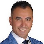 Bu, lacivert takım elbise giyen genç bir adamın görüntüsüdür. Adam öne doğru bakıyor ve ellerini önünde birleştirmiş. Bakımlı bir sakalı var ve yüzünde gözlük takıyor. Kahverengi saçları var ve takım elbisesi üzerine tam oturacak şekilde dikilmiş. Yakası hafifçe kalkık ve takım elbisesinin düğmeleri kapalı. Kendinden emin ve profesyonel görünüyor. Yüz ifadesi ciddi ve dik duruyor. 30'lu yaşlarının ortalarında görünüyor. Arka plan bulanık ve renkler soluk.