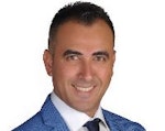 Bu, lacivert takım elbise giyen genç bir adamın görüntüsüdür. Adam öne doğru bakıyor ve ellerini önünde birleştirmiş. Bakımlı bir sakalı var ve yüzünde gözlük takıyor. Kahverengi saçları var ve takım elbisesi üzerine tam oturacak şekilde dikilmiş. Yakası hafifçe kalkık ve takım elbisesinin düğmeleri kapalı. Kendinden emin ve profesyonel görünüyor. Yüz ifadesi ciddi ve dik duruyor. 30'lu yaşlarının ortalarında görünüyor. Arka plan bulanık ve renkler soluk.