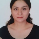Resimdeki kadının omuzlarının hemen altına düşen uzun siyah saçları var. Üzerinde V yakalı ve kısa kollu siyah bir tişört var. Kolları iki yanında ve nötr bir ifadeyle dümdüz karşıya bakıyor. İnce bir vücudu var ve arka planda beyaz bir duvarın olduğu aydınlık bir odada duruyor. Görüntüdeki ışık doğal, tenine sıcak ve sağlıklı bir parlaklık veriyor. Kameraya bakarken dudaklarında hafif bir gülümseme var.