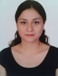 Resimdeki kadının omuzlarının hemen altına düşen uzun siyah saçları var. Üzerinde V yakalı ve kısa kollu siyah bir tişört var. Kolları iki yanında ve nötr bir ifadeyle dümdüz karşıya bakıyor. İnce bir vücudu var ve arka planda beyaz bir duvarın olduğu aydınlık bir odada duruyor. Görüntüdeki ışık doğal, tenine sıcak ve sağlıklı bir parlaklık veriyor. Kameraya bakarken dudaklarında hafif bir gülümseme var.