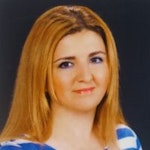 Uzun, siyah saçlı genç bir kadın mavi ve beyaz çizgili bir gömleğin önünde duruyor. Açık mavi renkteki gömleğin üzerinde hem yatay hem de dikey olarak uzanan beyaz çizgiler var. Saçları başının arkasında topuz yapılmış ve yüzü sol taraftan gelen sıcak bir ışıkla aydınlatılmış. Yüzünde sıcak ve huzurlu bir ifadeyle yana doğru bakıyor. Kollarını göğsünün önünde kavuşturmuş, kendinden emin ve güçlü bir duruş sergiliyor. Yirmili yaşlarının başında gibi görünüyor.