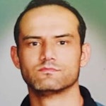 Koyu renk sakallı genç bir adam beyaz duvarlı bir odada durmaktadır. Yüzünde ciddi bir ifade var ve siyah bir tişört ile mavi bir kot pantolon giyiyor. Elleri yanında ve ayakları biraz açık. Kısa, koyu renk saçları ve güçlü bir çene çizgisi vardır. Sakalları kalın ve bakımlıdır. Gözleri derine dikilmiştir ve bakışları yoğundur. Uzun boylu ve gururludur, bir güç ve güven imajı çizer.