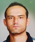Koyu renk sakallı genç bir adam beyaz duvarlı bir odada durmaktadır. Yüzünde ciddi bir ifade var ve siyah bir tişört ile mavi bir kot pantolon giyiyor. Elleri yanında ve ayakları biraz açık. Kısa, koyu renk saçları ve güçlü bir çene çizgisi vardır. Sakalları kalın ve bakımlıdır. Gözleri derine dikilmiştir ve bakışları yoğundur. Uzun boylu ve gururludur, bir güç ve güven imajı çizer.