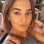Bu, selfie çeken genç bir kadının fotoğrafı. Arka planda parlak mavi gökyüzünün olduğu bir dış mekânda duruyor. Saçları açık kahverengi ve at kuyruğu şeklinde geriye doğru toplanmış. Beyaz bir tişört ve mavi kot pantolon giyiyor. Sol kolu dışarı doğru uzatılmış ve elinde bir akıllı telefon tutuyor. Gözleri kapalı ve gülümsüyor. Memnun ve rahat görünüyor. Bir ayağını hafifçe yerden kaldırmış, bu da onu eğlenceli bir ruh hali içinde gösteriyor.
