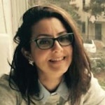 Gözlük takan ve genişçe gülümseyen genç bir kadın. Orta uzunlukta kahverengi saçları var ve gözleri parlak ve ışıltılı. Gülümsemesi geniş ve doğrudan kameraya bakıyor. Pembe yanakları ve dostça bir ifadesi var. Gözlükleri kahverenginin açık bir tonunda ve yüzünde hafifçe orantısız duruyor. Üzerinde gri bir gömlek ve boynunda siyah bir eşarp var. Rahat ve mutlu görünüyor.