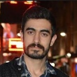 Orta yaşlı, gür sakallı ve bıyıklı bir adam kameranın önünde duruyor. Yüzünde sert bir ifade, güçlü bir çene hattı ve belirgin elmacık kemikleri vardır. Siyah bir tişört ve kot pantolon giymektedir ve erkeksi bir yapısı vardır. Sakalı ve bıyığı bakımlıdır ve saçları düzgünce geriye taranmıştır. Kendinden emin ve kararlı görünüyor, bir güç ve erkeklik sembolü.