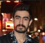 Orta yaşlı, gür sakallı ve bıyıklı bir adam kameranın önünde duruyor. Yüzünde sert bir ifade, güçlü bir çene hattı ve belirgin elmacık kemikleri vardır. Siyah bir tişört ve kot pantolon giymektedir ve erkeksi bir yapısı vardır. Sakalı ve bıyığı bakımlıdır ve saçları düzgünce geriye taranmıştır. Kendinden emin ve kararlı görünüyor, bir güç ve erkeklik sembolü.