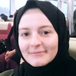 Görüntüdeki kadın koyu tenli ve başını ve omuzlarını örten siyah bir başörtüsü takıyor. Saçları geriye çekilmiş ve toplanmış. Düşünceli bir ifadeyle kameradan uzağa bakıyor. Gözleri kahverengi ve ağzı hafifçe açık. Üzerinde hafif desenli beyaz bir üstlük ve tek boncuklu bir kolye var. Boynundan aşağı sarkan uzun küpeleri var ve kollarını önünde kavuşturmuş. Kendine güvenen ve dengeli bir duruşu var.