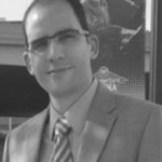 Bu, açık mavi kravatlı, şık, lacivert bir takım elbise giyen bir adamın fotoğrafı. Düzgünce şekillendirilmiş kısa siyah saçları ve güçlü bir çene hattı var. Kendine güvenen ve dengeli bir ifadeyle doğrudan kameraya bakıyor. Duruşu düzgün ve elleri önünde kenetlenmiş. Beyaz bir gömlek, siyah elbise ayakkabıları ve gümüş bir saat giyiyor. Yirmili yaşlarının sonunda ya da otuzlu yaşlarının başında görünüyor.
